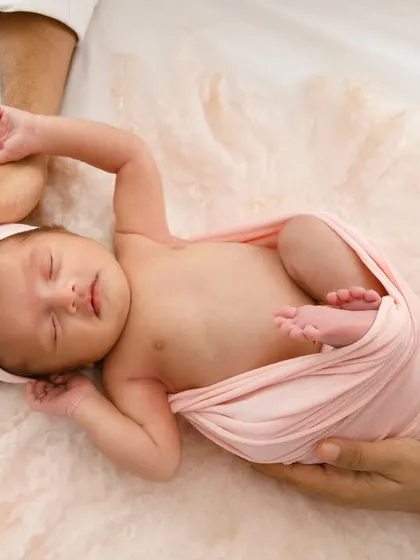 Held safely in their parents' hands, this sleeping baby girl is a picture of serenity. The soft pink wrap and headband add a delicate touch to this newborn portrait.