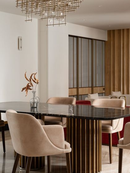 The dining room at Haus of Aureus features a black marble tabletop with a sculptural brass and wood base. The plush, beige velvet chairs and a modern brass chandelier complete this luxurious and sophisticated entertaining space.