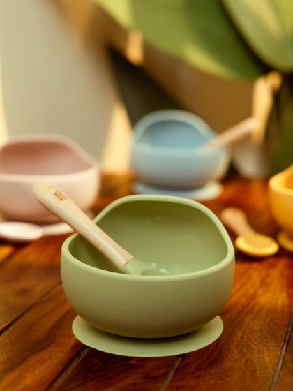 A close-up of silicone baby bowls in various colors, shot in a warm, home-like setting. The soft lighting gives a gentle and safe feel.