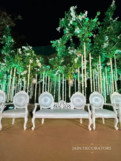 Vintage-style seating is arranged before an enchanted forest backdrop. The ornate white chairs provide an elegant contrast to the organic, whimsical design of the illuminated trees and hanging florals.