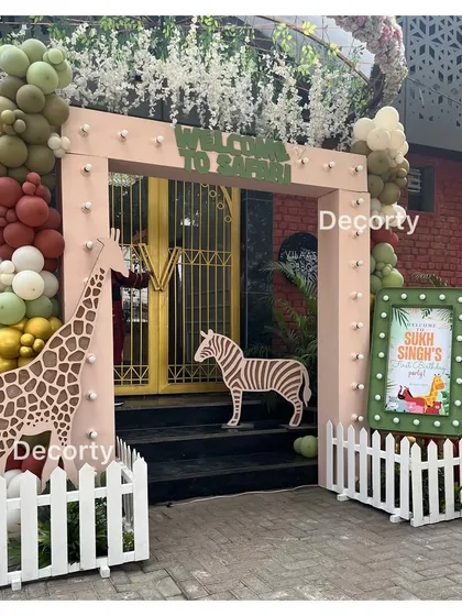 A fun jungle safari entrance with giraffe and zebra cutouts, a white picket fence, and earthy-toned balloons.