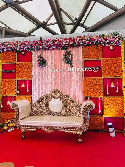 A festive stage with a backdrop of marigold checkerboard panels and a curtain of white flowers. The ornate sofa and hanging bells complete this traditional look for an engagement or sangeet.