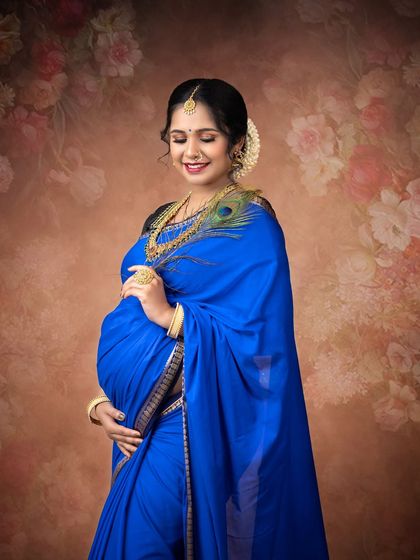 A lovely solo portrait of the mother-to-be in a vibrant blue saree, holding a peacock feather. The floral background adds a soft, feminine touch to this traditional look.