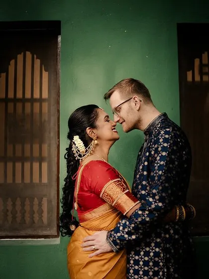 A cross-cultural couple shares an intimate moment against a green wall, their contrasting outfits telling a story of two worlds meeting.