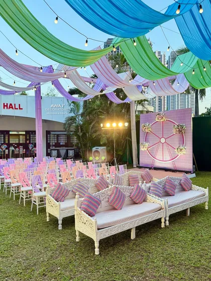A whimsical outdoor Mehendi or Haldi setup featuring pastel-colored drapes, traditional carved white seating, and a unique Ferris wheel backdrop.