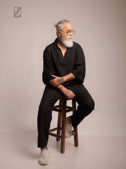 This seated profile shot emphasizes the model's distinctive look, including his hair and beard, against a clean studio background.