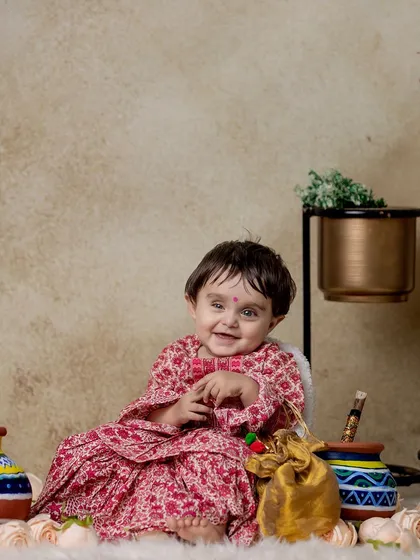 A full-length portrait of a baby girl in a lovely printed dress for her Radha-themed shoot, surrounded by props in a comfortable home setup.