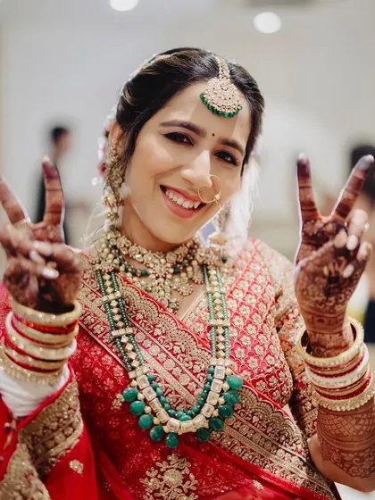Peace, love, and bridal glam! This happy bride is all set for her phera ceremony, glowing in a traditional red lehenga with soft makeup and elegant emerald jewelry.