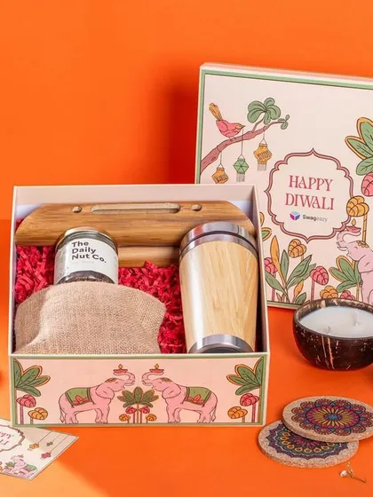A Diwali hamper from Swageasy, styled against a bright orange background. The box contains a mix of modern and traditional items, like a bamboo tumbler and dry fruits, appealing to a corporate gifting audience.