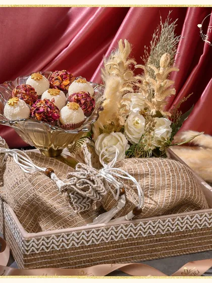 Elegance wrapped in tradition. This luxurious hamper features handcrafted delicacies in rustic jute potlis, arranged in a beautiful woven tray with dried floral accents for a chic, earthy feel.