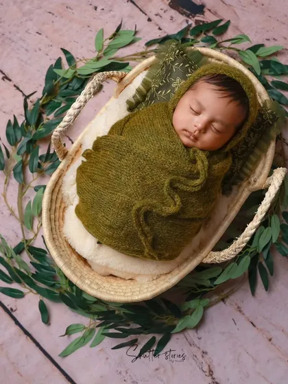 A newborn wrapped in an olive green knit outfit and bonnet, resting in a basket surrounded by green leaves.