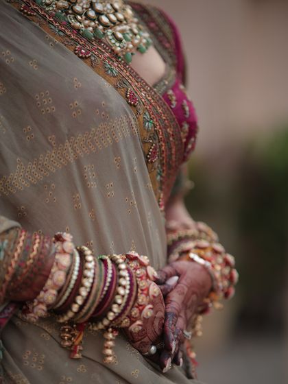 A detail shot of the bride's hands, adorned with mehendi and a stunning stack of traditional wedding bangles. The texture and colors of her lehenga and blouse are also highlighted here.