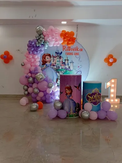 A princess-themed first birthday party featuring Sofia the First. The decor includes a circular backdrop with the birthday girl's name, character cutouts, themed pedestals, and a purple and pink balloon garland.