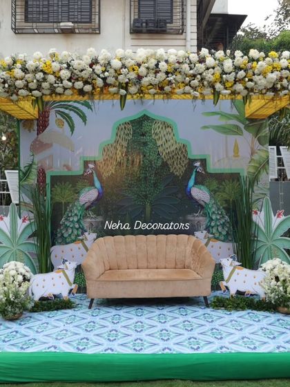 A whimsical and artistic Mehendi stage. The backdrop features a custom illustration of peacocks in a lush garden, complemented by a comfortable sofa and unique animal props for a touch of fun.
