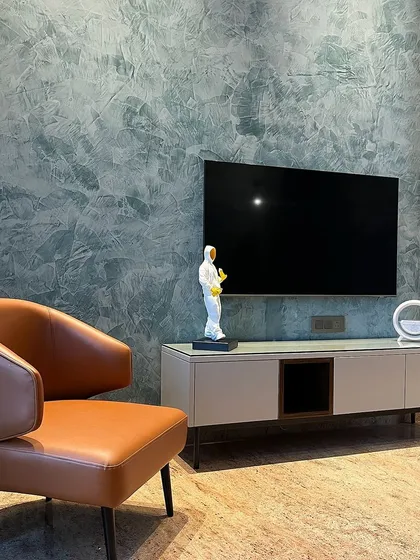 A close-up of a TV corner in an Ambala bedroom. The textured green wall paint provides a dramatic backdrop for the minimalist console and a tan leather accent chair, creating a stylish contrast.