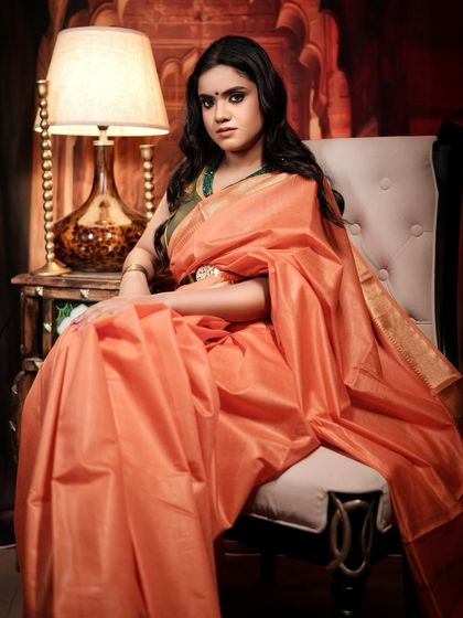 A full-length seated portrait that displays the beautiful drape of the orange silk saree and the elegance of the pose.