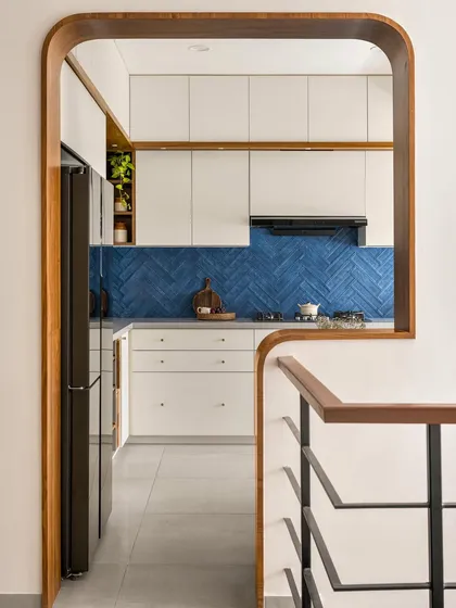 The entryway to the 'Palaash' kitchen is framed by a custom curved wood arch, offering a glimpse of the blue herringbone tiles and clean white cabinetry within.