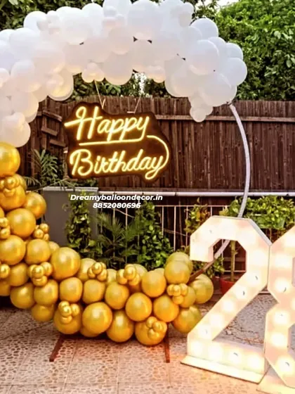 A chic outdoor birthday setup for a 28th birthday. The half-arch of gold and white balloons looks like clouds, paired with a neon sign and large marquee numbers.