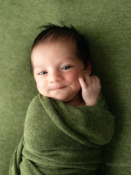A newborn gives a sweet, sly smile while wrapped in a green swaddle. The soft, natural light beautifully highlights the curve of his smile and delicate features.