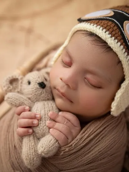 A close up of our tiny pilot, cuddling a teddy bear. This shot focuses on the baby's peaceful expression and the adorable details of the hand knit aviator hat.