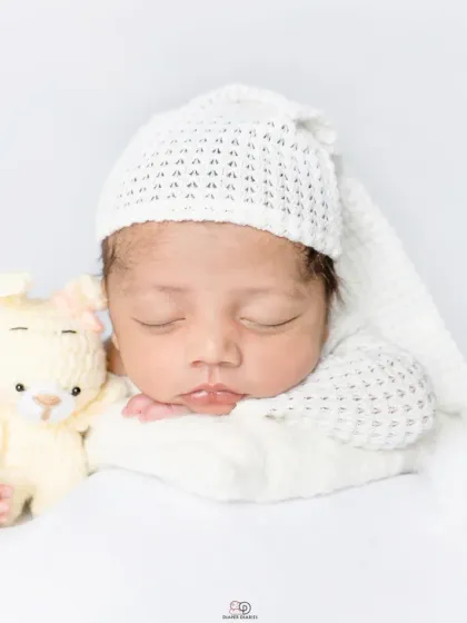 A pure and simple newborn portrait against a clean white background. The baby is dressed in a white knitted outfit and sleepy hat, showcasing their angelic features.