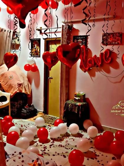 A cozy room filled with red and white balloons for a romantic surprise. The decor features 'Love' and heart-shaped foil balloons, with petals scattered across the floor for an intimate feel.