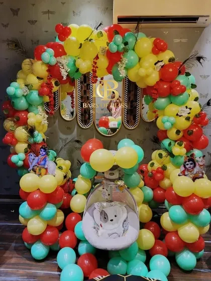 A vibrant and traditional Janmashtami decor for a baby's first festival. The setup includes a colorful balloon arch and a decorated swing (jhula) for the baby, adorned with peacock feathers.