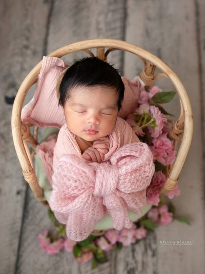 A nine-day-old newborn girl wrapped in a pink bow, resting in a tiny wicker chair. The composition is sweet, delicate, and full of charm.