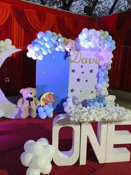 A dreamy 'teddy bear on the moon' theme for a first birthday. The decor features a large 'ONE' table, a moon prop, and a blue and white color scheme with fluffy clouds.