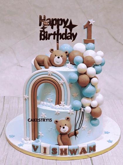 A dreamy two-tier teddy bear cake for a first birthday. This 4kg chocolate cake features a beautiful blue color, fondant balloon cascades, a rainbow, and cute teddy figures.
