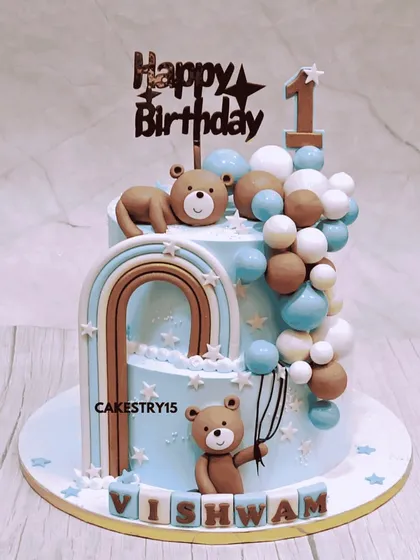 A dreamy two-tier teddy bear cake for a first birthday. This 4kg chocolate cake features a beautiful blue color, fondant balloon cascades, a rainbow, and cute teddy figures.