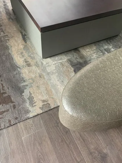 A detail showing the intersection of different textures: a patterned rug, a smooth wooden floor, and an embossed leather stool.