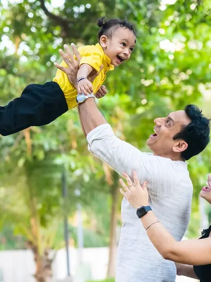 A joyful, candid moment captured outdoors as a father playfully lifts his laughing child high into the air, with the mother looking on with a smile.