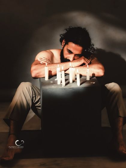 A portrait of weariness or deep thought, resting his head near the candles. The image is raw, emotional, and full of unspoken narrative.
