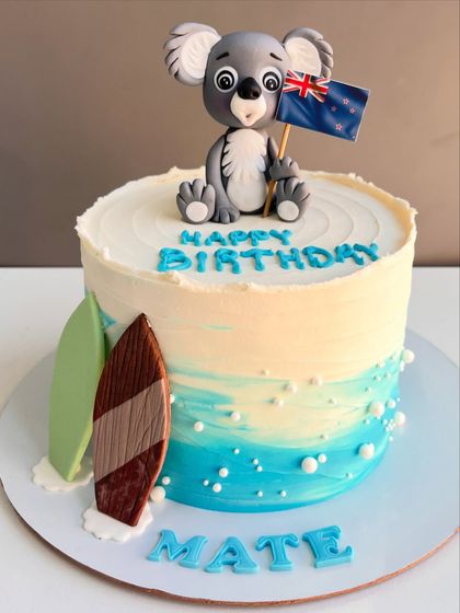 This cake takes you straight to the beach with its ocean-hued frosting and sandy details. It's topped with a cute, hand-sculpted koala holding a New Zealand flag and features two surfboards, perfect for a "Happy Birthday Mate" celebration.