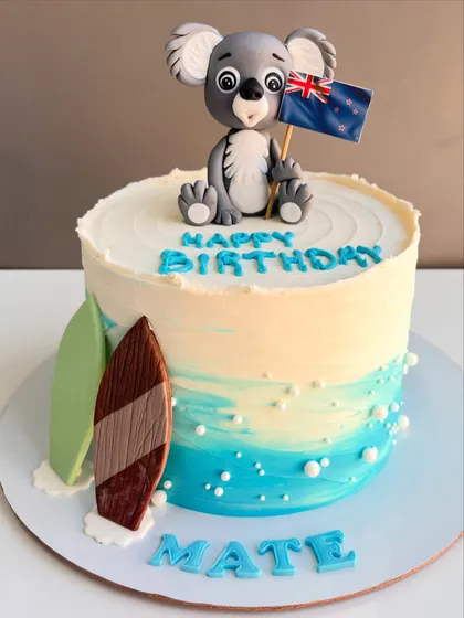 This cake takes you straight to the beach with its ocean-hued frosting and sandy details. It's topped with a cute, hand-sculpted koala holding a New Zealand flag and features two surfboards, perfect for a "Happy Birthday Mate" celebration.
