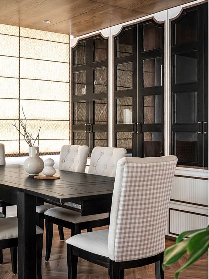 The formal dining room at Riviera Maison, featuring a large dark wood table and a mix of tufted and gingham-upholstered chairs. The black glass-front cabinets add a sense of drama and scale to the space.