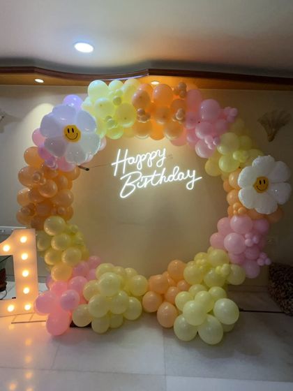 A sweet and sunny first birthday setup with a daisy theme. The circular balloon arch in pastel orange, yellow, and pink is decorated with large, smiling daisy balloons. The light-up number '1' stands proudly beside it.