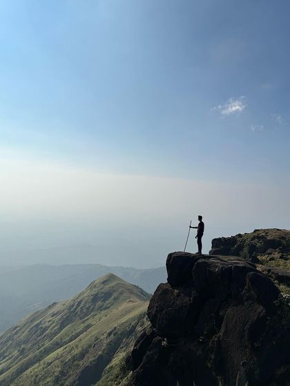 A trekker with a walking stick stands at the edge of a cliff on Kumara Parvatha, overlooking the stunning Western Ghats.