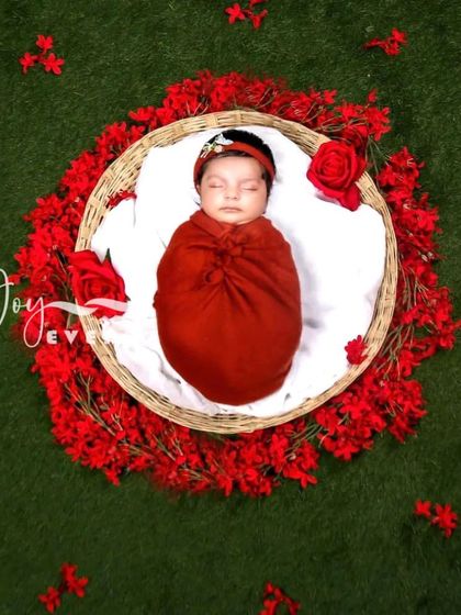 This overhead shot captures a newborn swaddled in red, placed in a basket surrounded by a ring of bright red flowers against a green grass backdrop. The color contrast makes this a visually striking image.