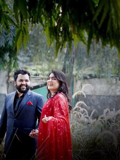 A joyful, candid shot of a couple walking through a garden. The woman's bright red saree and genuine laughter make this a vibrant and happy portrait.