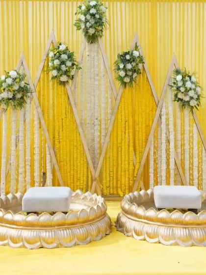 Here is the main stage for the Haldi ritual, featuring two traditional bajots. The backdrop is a geometric wooden frame decorated with marigold and tuberose strings, blending modern design with traditional flowers.