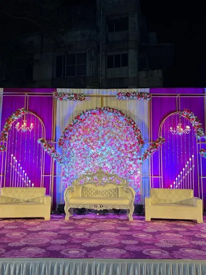 This reception stage is designed with a rich purple and white theme. The backdrop features arched panels, chandeliers, and a dense floral centerpiece, all coordinated with the patterned purple carpet.