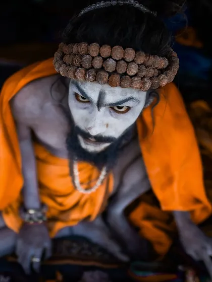 A high-angle portrait of a Naga sadhu with piercing eyes, looking up at the camera. The orange robe and ash-smeared skin create a stark and memorable image of asceticism.