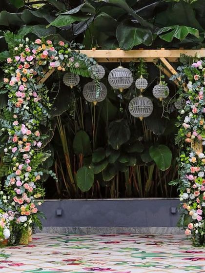 A close-up of a floral arch against a wall of lush greenery. The mix of pastel flowers and hanging lanterns is perfect for a romantic reception or engagement party.