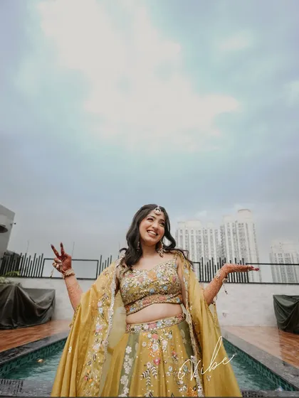 A happy bride strikes a pose by the pool during her Mehendi function. The low-angle shot against the cloudy sky creates a dramatic and joyful portrait.