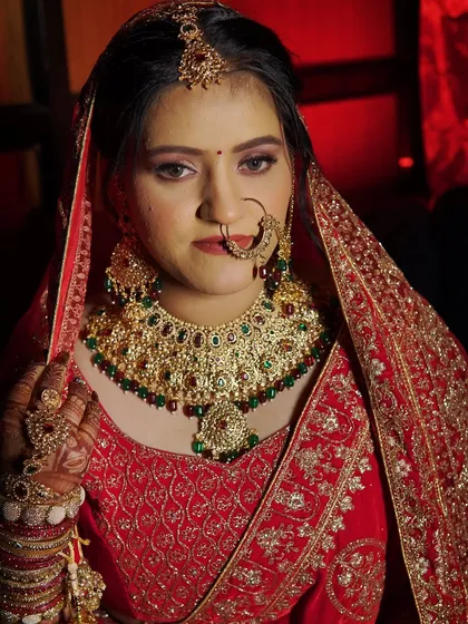 A close-up portrait against a dramatic red background. The lighting highlights the beautiful details of her makeup and the stunning bridal jewelry.