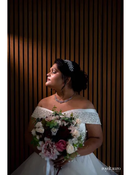 A beautiful color portrait of a bride in her wedding gown, holding her bouquet. A sliver of light illuminates her face, creating a serene and graceful image against the modern, textured wall.