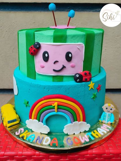 A fun, two-tier Cocomelon cake where the top tier is shaped like the iconic melon TV. The cake is decorated with a rainbow, ladybugs, and a little JJ figurine.