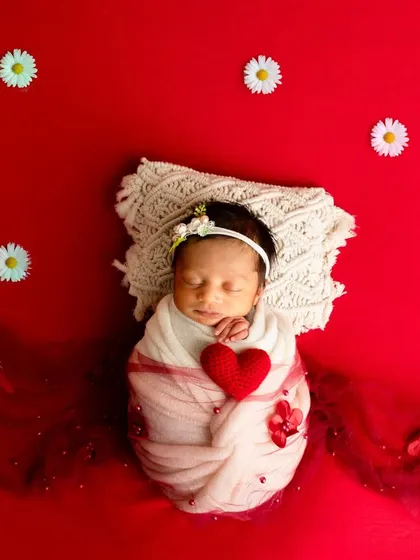 A newborn swaddled in white holds a red heart, lying on a red background dotted with white daisies for a classic, high-contrast look.